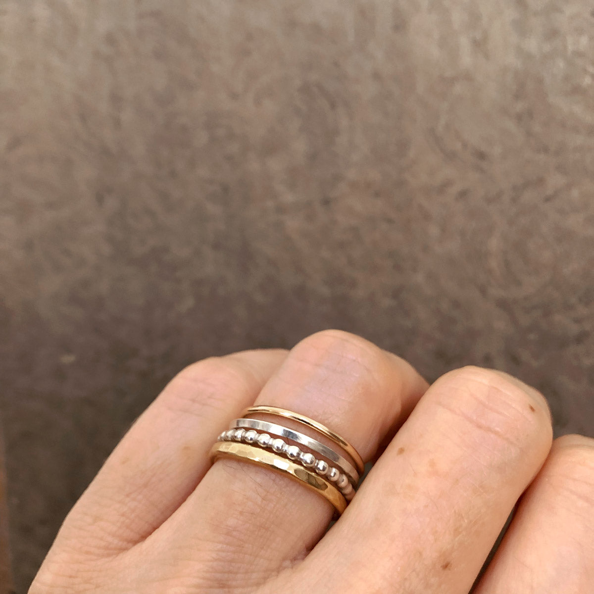Top view of Alpine Loop stacking ring set worn on a finger—four handcrafted mixed metal rings including hammered brass, beaded sterling silver, square silver, and gold-filled bands by Narrow-Gauge Designs