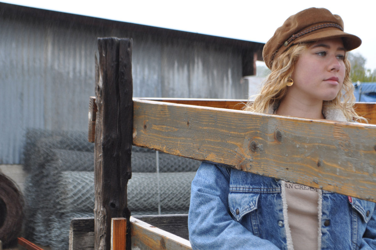 Model wearing Billy the Kid gold minimalist earrings, denim jacket, and hat, standing in rustic fenced-in area, evoking a relaxed Old West vibe.