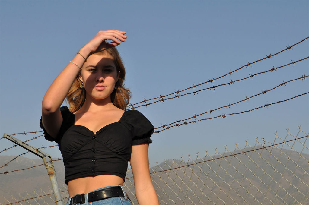 A blonde model is shielding her eyes from the sun as she poses in front of a barbed wire fence and Palomar Mountain.