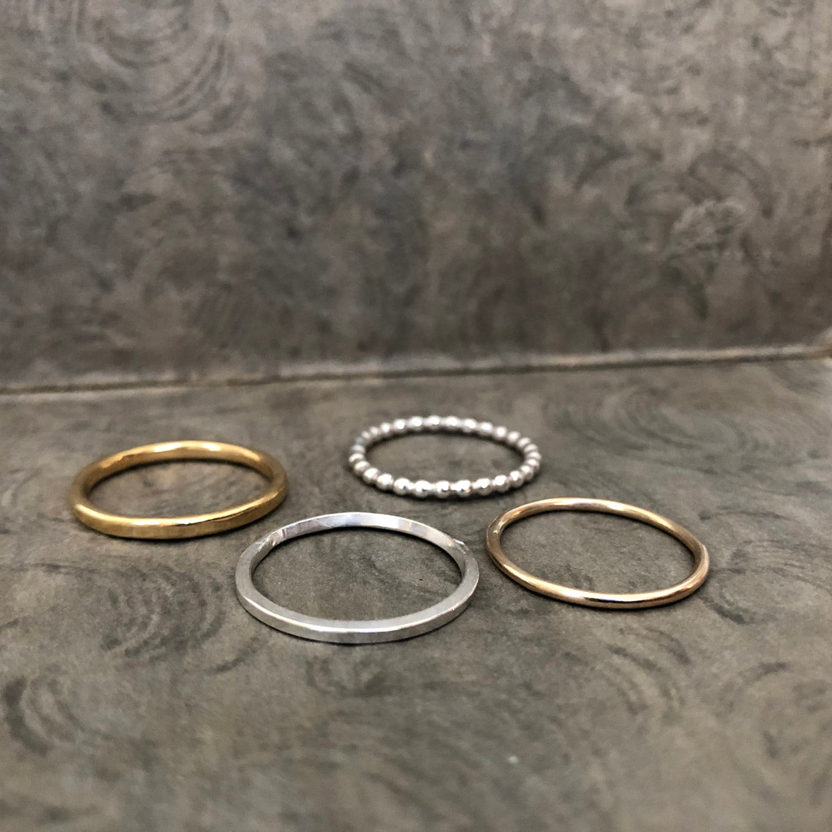 Four handcrafted stacking rings from the Alpine Loop set lying flat—hammered brass, beaded sterling silver, square silver, and gold-filled bands, by Narrow-Gauge Designs