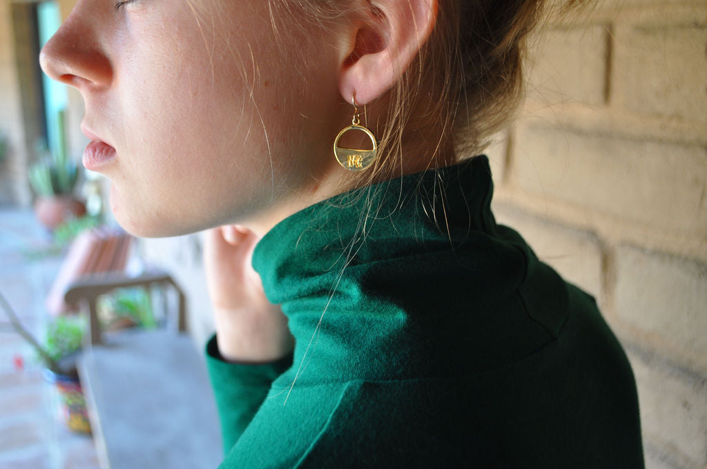 Profile shot of young woman wearing Billy the Kid gold minimalist earrings with hair up and green turtleneck; Narrow-Gauge logo clearly visible on earring.