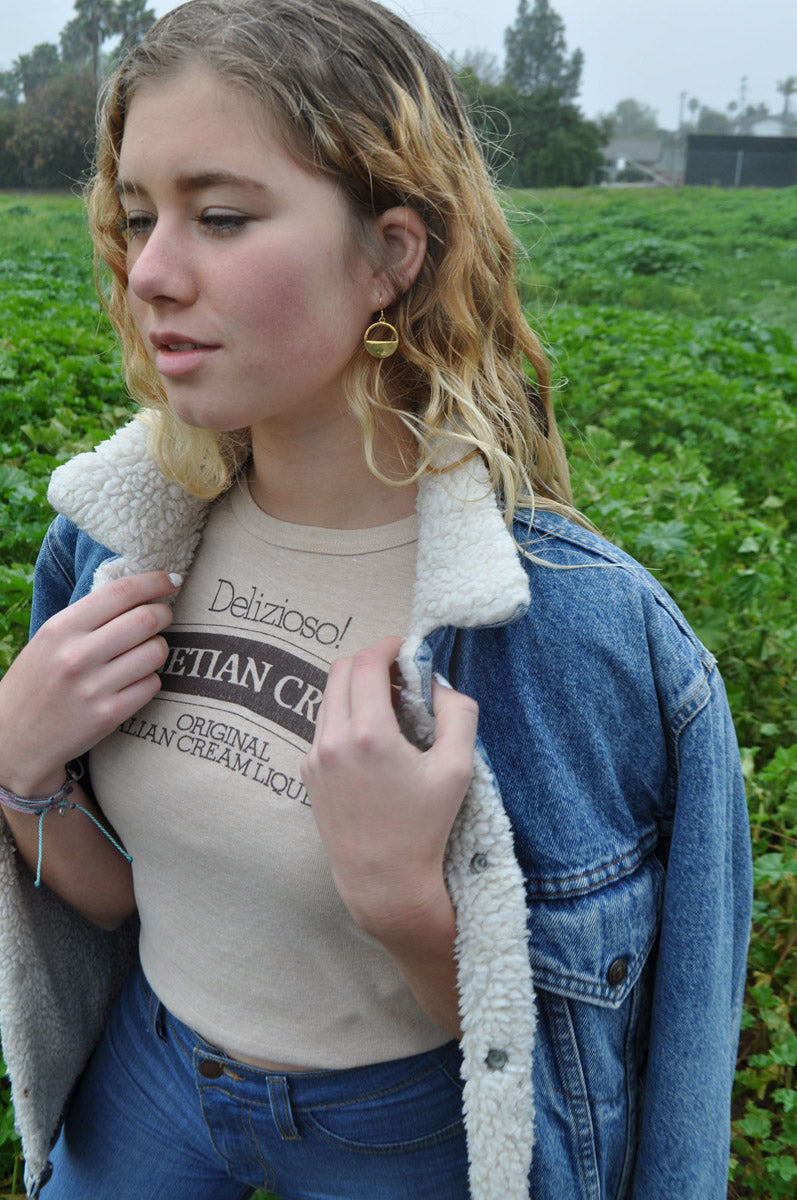 Model wearing Billy the Kid gold minimalist earrings and denim jacket, standing in tall weeds, showcasing the earrings’ subtle shine and casual style.