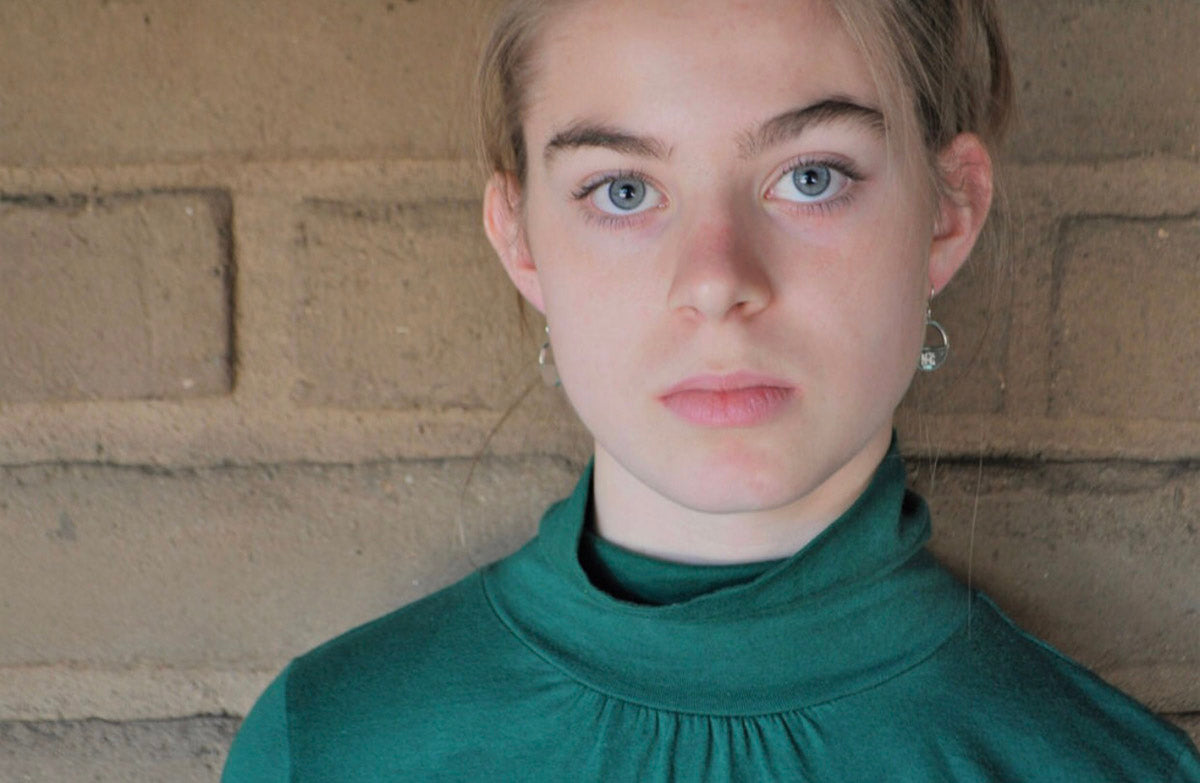 Young model wearing Billy the Kid sterling silver minimalist earrings and green turtleneck, gazing directly into the camera with a confident expression.