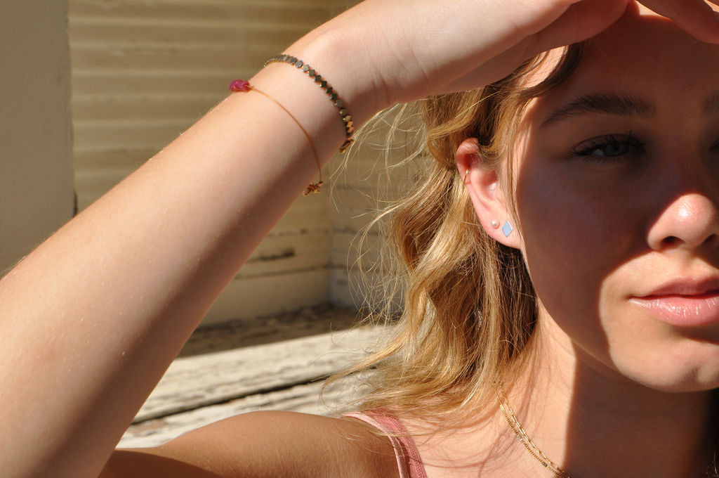 Model wearing diamond-shaped Pharo sterling silver earring and Narrow-Gauge bracelets, shielding eyes from the sun while dressed in a light-colored outfit.